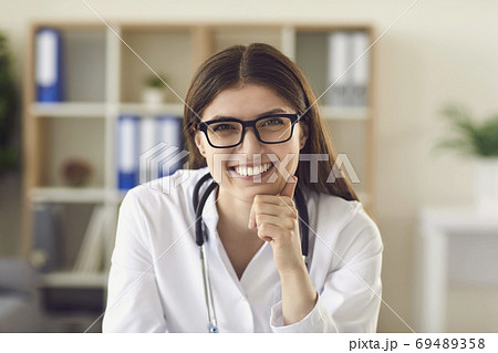 Portrait of friendly young female physician at desk in office smiling to patient Portrait of friendly young female physician at desk in office smiling to patient 69489358