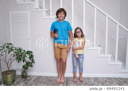 Homeschooling. Brother and sister standing near stairs at home, looking at camera and smiling after studying online or doing homework together Homeschooling. Brother and sister standing near stairs at home, looking at camera and smiling after studying online or doing homework together 69491241