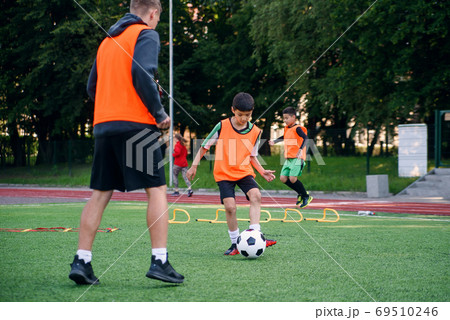 Skilled child player in football uniform working out the kicking ball together with his experienced coach on sport field Skilled child player in football uniform working out the kicking ball together with his experienced coach on sport field 69510246