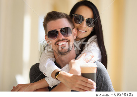 Beautiful woman in sunglasses hug man behind neck and hold cardboard glass with coffee in her hand. 69540305