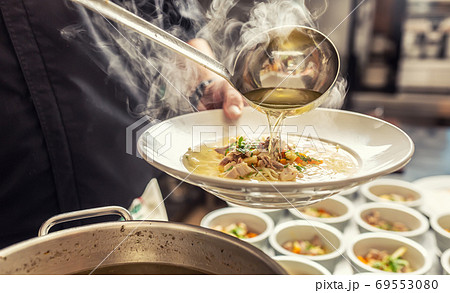 Chef pours broth into a chicken soup with noodles, meat and vegetable 69553080