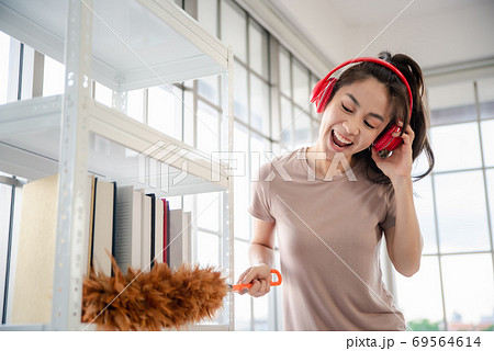 Young asian woman with headphones using a feather duster to clean wood book shelves in home . 69564614