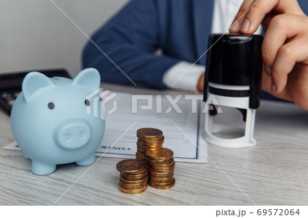Businessman is stamping contract in office. Blue piggy bank with stack of coins on desk. Economy and management financial concept 69572064
