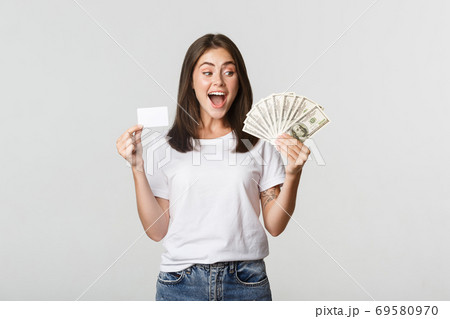 Amused beautiful girl looking at money and holding credit card, standing white background 69580970