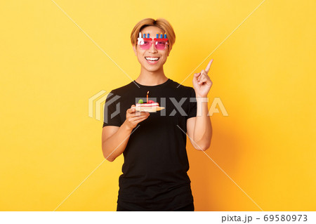 Cheerful, handsome asian birthday guy in funny sunglasses, holding b-day cake with candle and pointing finger upper right corner at your logo, yellow background 69580973