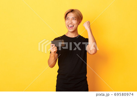 Portrait of stylish handsome asian guy playing smartphone game, rejoicing from winning, standing yellow background Portrait of stylish handsome asian guy playing smartphone game, rejoicing from winning, standing yellow background 69580974