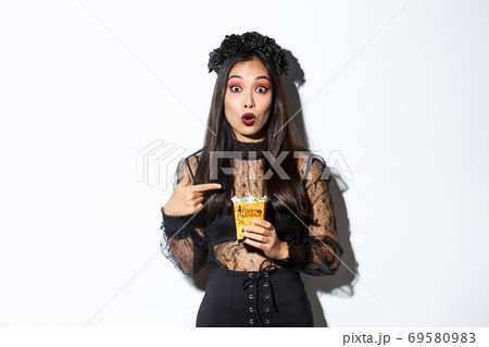 Amazed asian girl looking at camera while pointing finger at sweets, gather treats on halloween, wearing witch dress, standing over white background Amazed asian girl looking at camera while pointing finger at sweets, gather treats on halloween, wearing witch dress, standing over white background 69580983