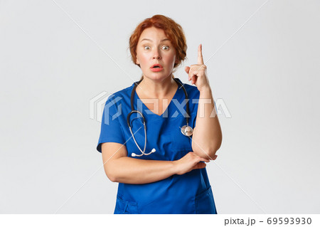 Overwhelmed redhead female doctor, middle-aged nurse in scrubs have suggestion, saying idea or plan, raising index finger eureka gesture with worried expression, standing grey background Overwhelmed redhead female doctor, middle-aged nurse in scrubs have suggestion, saying idea or plan, raising index finger eureka gesture with worried expression, standing grey background 69593930