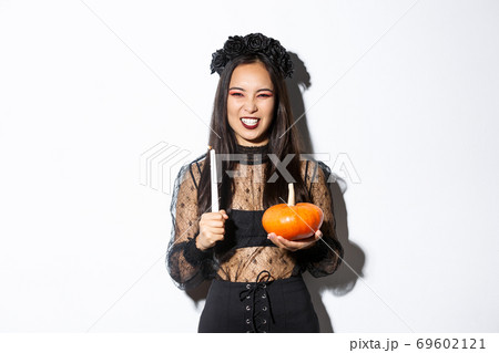 Image of asian evil witch in gothic lace dress and black wreath, laughing and grimacing, holding candle with pumpkin, celebrating halloween 69602121