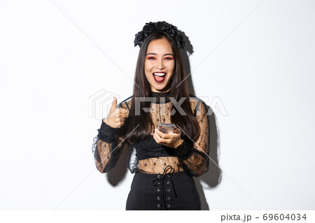 Portrait of satisfied asian woman in elegant gothic dress and black wreath showing thumbs-up while using mobile phone, standing over white background Portrait of satisfied asian woman in elegant gothic dress and black wreath showing thumbs-up while using mobile phone, standing over white background 69604034
