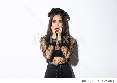Shocked young asian woman wearing witch costume, looking concerned. Female in black dress and wreath looking like widow or magician gasping worried, standing white background 69604063