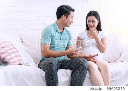 Husband holding glass of fresh water take care his wife pregnant sitting on white sofa 69605279