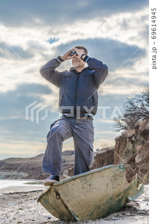 man stands in a broken boat on the beach and carefully looks through binoculars 69614945