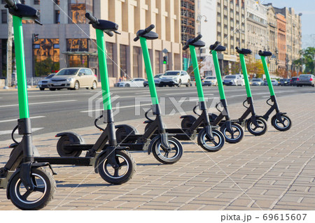 Eco transportation concept. Scooter rental in the city. Environment conservation. Eco friendly transportation. Vehicle rent service concept. Electric scooters in line on urban city background Eco transportation concept. Scooter rental in the city. Environment conservation. Eco friendly transportation. Vehicle rent service concept. Electric scooters in line on urban city background 69615607