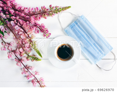 Cup of coffee, removed medical mask and pink flowers. Breakfast in the cafe. Modern reality and life concept. Blue surgical protective mask and white cup on white wooden background. Flat lay, top view Cup of coffee, removed medical mask and pink flowers. Breakfast in the cafe. Modern reality and life concept. Blue surgical protective mask and white cup on white wooden background. Flat lay, top view 69626970
