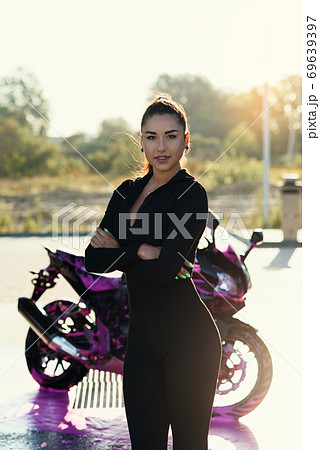 Seductive young woman in tight fitting black suit poses near sport motorcycle at self service car wash. Seductive young woman in tight fitting black suit poses near sport motorcycle at self service car wash. 69639397