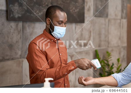 African male receptionist wearing medical mask using infrared thermometer for measuring body temperature of office worker 69644501