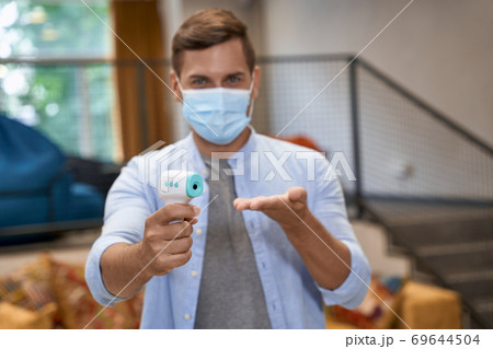 Business during Covid 19 or coronavirus, portrait of young male office worker wearing medical protective mask standing in the office and showing infrared thermometer at camera Business during Covid 19 or coronavirus, portrait of young male office worker wearing medical protective mask standing in the office and showing infrared thermometer at camera 69644504