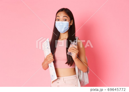 Safe tourism, travelling during coronavirus pandemic and preventing virus concept. Cute asian girl tourist on vacation with medical mask and hand sanitizer, holding map of city, pink background 69645772