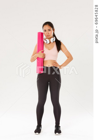 Full length of gloomy and upset cute brunette asian fitness girl in sportswear, sulking while holding rubber mat for yoga classes, looking disappointed as gym closed during covid-19 quarantine 69645828