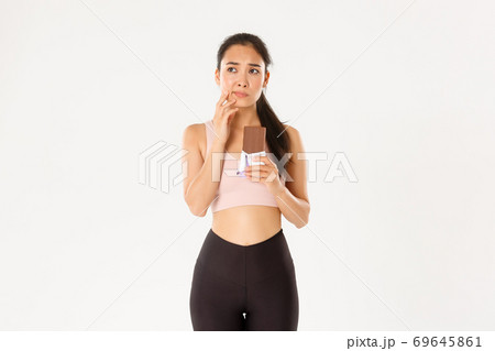 Sport, wellbeing and active lifestyle concept. Doubtful and indecisive asian fitness girl holding chocolate bar and thinking about eating it, looking away hesitant, worried about calories 69645861