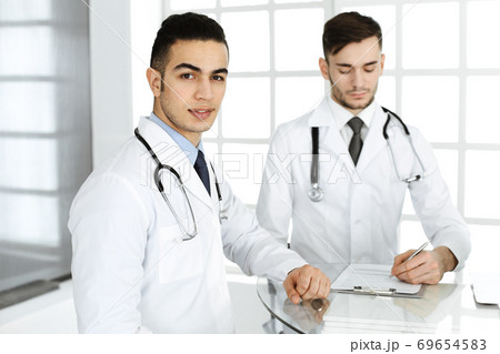 Arab doctor man sitting at the glass desk with caucasian colleague in medical office or clinic. Diverse doctors team, medicine and healthcare concept 69654583