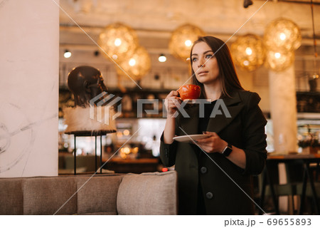 Beautiful young woman holding cup of tasty hot coffee in hand while standing in restaurant. Beautiful young woman holding cup of tasty hot coffee in hand while standing in restaurant. 69655893