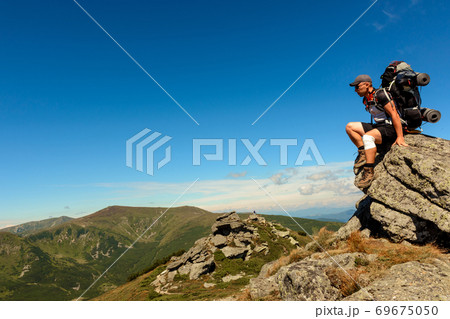 Young man with a backpack standing on top of a mountain and enjoying the view of the valley, the top of the Carpathians Eared stone. Young man with a backpack standing on top of a mountain and enjoying the view of the valley, the top of the Carpathians Eared stone. 69675050