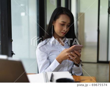 Female texting on smartphone while sitting at worktable in office room Female texting on smartphone while sitting at worktable in office room 69675574