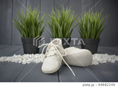 Newborn baby boy's booties in white, with decorative stones, green plants and a gray wooden background. Children's shoes 69682240