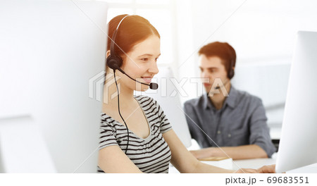 Casual dressed young woman using headset and computer while talking with customers online. Group of operators at work in sunny office. Call center 69683551