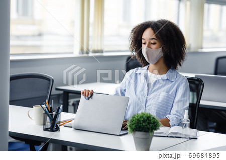 End of day and go home. African american woman in protective mask closes laptop, sitting at table with antiseptic in modern office interior End of day and go home. African american woman in protective mask closes laptop, sitting at table with antiseptic in modern office interior 69684895
