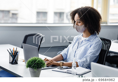 New normal and office work with protective mask and antiseptic. African american woman typing on laptop with notepad to write customer orders in modern interior 69684962
