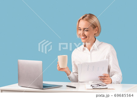 Positive business lady working with documents and drinking coffee in front of laptop computer at desk, blue background Positive business lady working with documents and drinking coffee in front of laptop computer at desk, blue background 69685564