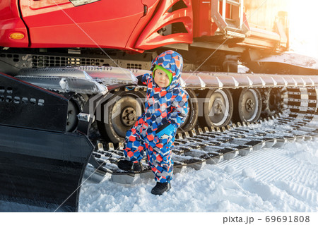 Cute adorable playful happy toddler boy inside red modern snowcat ratrack snowplow box grooming standing on peak alpine skiing resort Ischgl Austria. Heavy machinery mountain equipment track vehicle Cute adorable playful happy toddler boy inside red modern snowcat ratrack snowplow box grooming standing on peak alpine skiing resort Ischgl Austria. Heavy machinery mountain equipment track vehicle 69691808