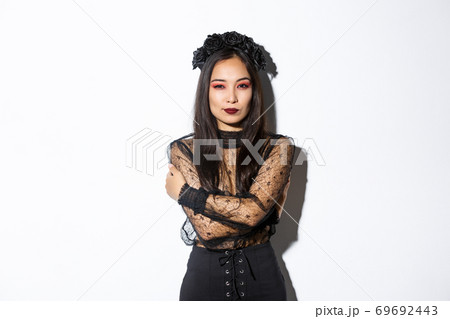 Young elegant woman in stylish gothic dress and wreath, embracing her body and looking sassy at camera, wearing halloween costume of undead widow, standing over white background Young elegant woman in stylish gothic dress and wreath, embracing her body and looking sassy at camera, wearing halloween costume of undead widow, standing over white background 69692443