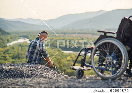 Military disabled man sitting on the mountain near wheelchair and raised hands up enjoying fresh air and nature beauty. Military disabled man sitting on the mountain near wheelchair and raised hands up enjoying fresh air and nature beauty. 69694376