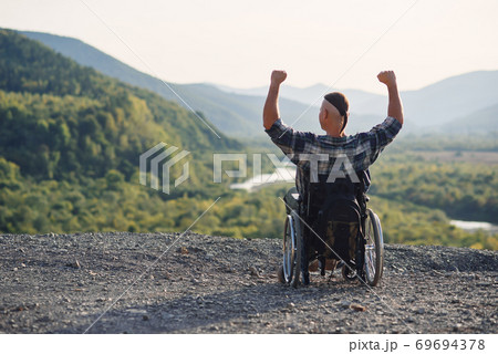 Lonely young man in a wheelchair enjoying fresh air in sunny day on the mountain. People with disabilities travelling. Lonely young man in a wheelchair enjoying fresh air in sunny day on the mountain. People with disabilities travelling. 69694378