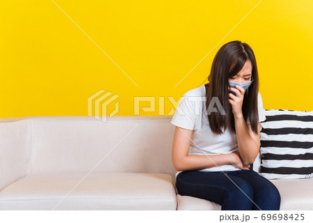 Woman sitting on sofa wearing protection face mask against coronavirus her sneeze hand close mouth Woman sitting on sofa wearing protection face mask against coronavirus her sneeze hand close mouth 69698425