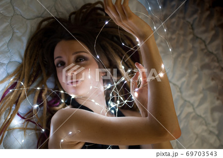 Indoors female portrait of a woman with dreadlocks hairstyle and smoky eyes makeup holding lights garland near her face, femininity and hollidays concepts 69703543