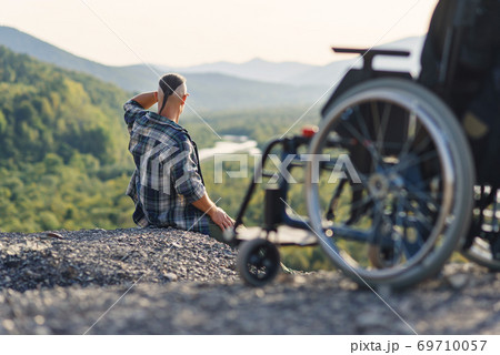 Disabled man sits on the mountain top near wheelchair and enjoying life and nature beauty. Selective focus. 69710057