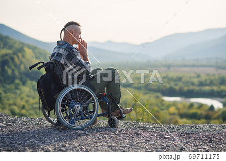 Young pensive man in a wheelchair enjoying the beauty of nature in the mountains. Rehabilitation after injury. 69711175