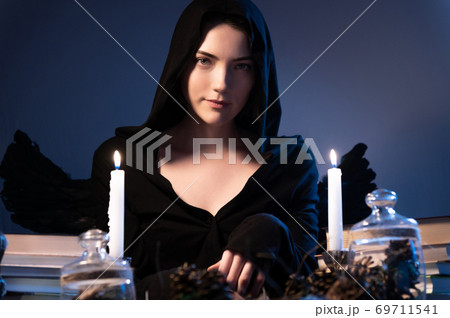 Medium closeup attractive Caucasian girl with golden long hair in a black hood sits at a table and leafs through a book. Costume with wings. Candles are burning. Halloween celebration concept Medium closeup attractive Caucasian girl with golden long hair in a black hood sits at a table and leafs through a book. Costume with wings. Candles are burning. Halloween celebration concept 69711541