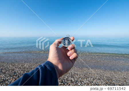 Compass in hand at seacoastA man's hand holds a magnetic compass against the background of a calm morning sea with haze above the water surface Compass in hand at seacoastA man's hand holds a magnetic compass against the background of a calm morning sea with haze above the water surface 69711542