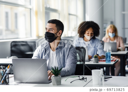 New rules for working in office and create a new idea. Arabian guy in protective mask looks out the window and works at laptop at workplace with antiseptic New rules for working in office and create a new idea. Arabian guy in protective mask looks out the window and works at laptop at workplace with antiseptic 69713825