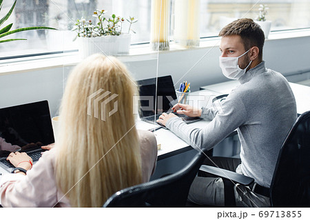 Social distance and working in modern office. Colleagues are guy and lady in protective masks talk through protective glass during COVID-19 pandemic Social distance and working in modern office. Colleagues are guy and lady in protective masks talk through protective glass during COVID-19 pandemic 69713855