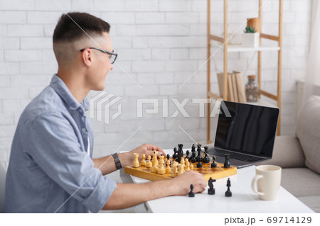 Smart guy and hobby in free time on self-isolation. Happy chess player in glasses with chess board looking at laptop with blank screen in living room interior 69714129