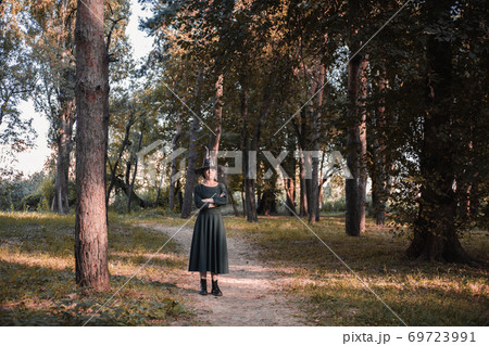 Pretty young beautiful woman in dark dress and witch hat standing in middle of autumn woods or park. Halloween party costume. Portrait in full growth. Shadow on the face. 69723991
