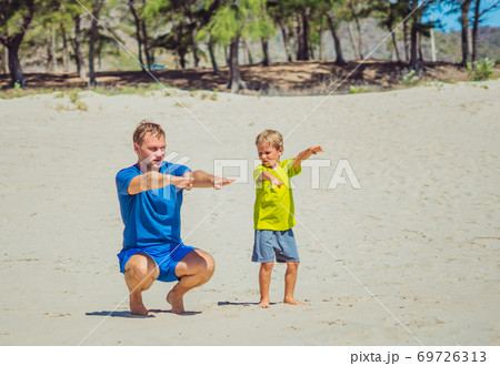 Sport for little children. Father cute son exercising on sand beach near forest park, having workout enjoying together in the fresh air, correct squatting technique, daddy influence on boy worldview Sport for little children. Father cute son exercising on sand beach near forest park, having workout enjoying together in the fresh air, correct squatting technique, daddy influence on boy worldview 69726313