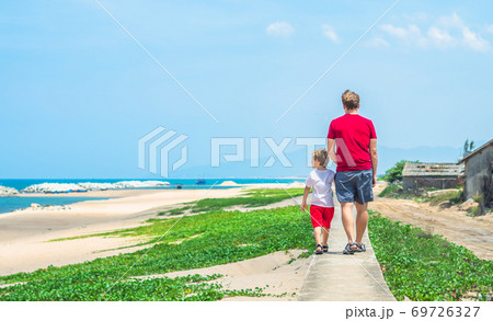 Father son walk along narrow concrete path, sea blue clear sky green grass background. Symbol as dad leads child into future adulthood. Happy childhood, fathers day, daddy influence on boy worldview 69726327